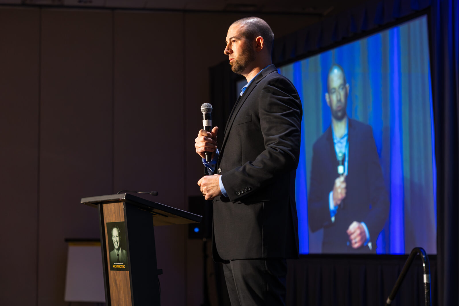 Dan Coomes-Barry presenting from the lectern at AGOGE 2024, his image projected on the screen behind him.