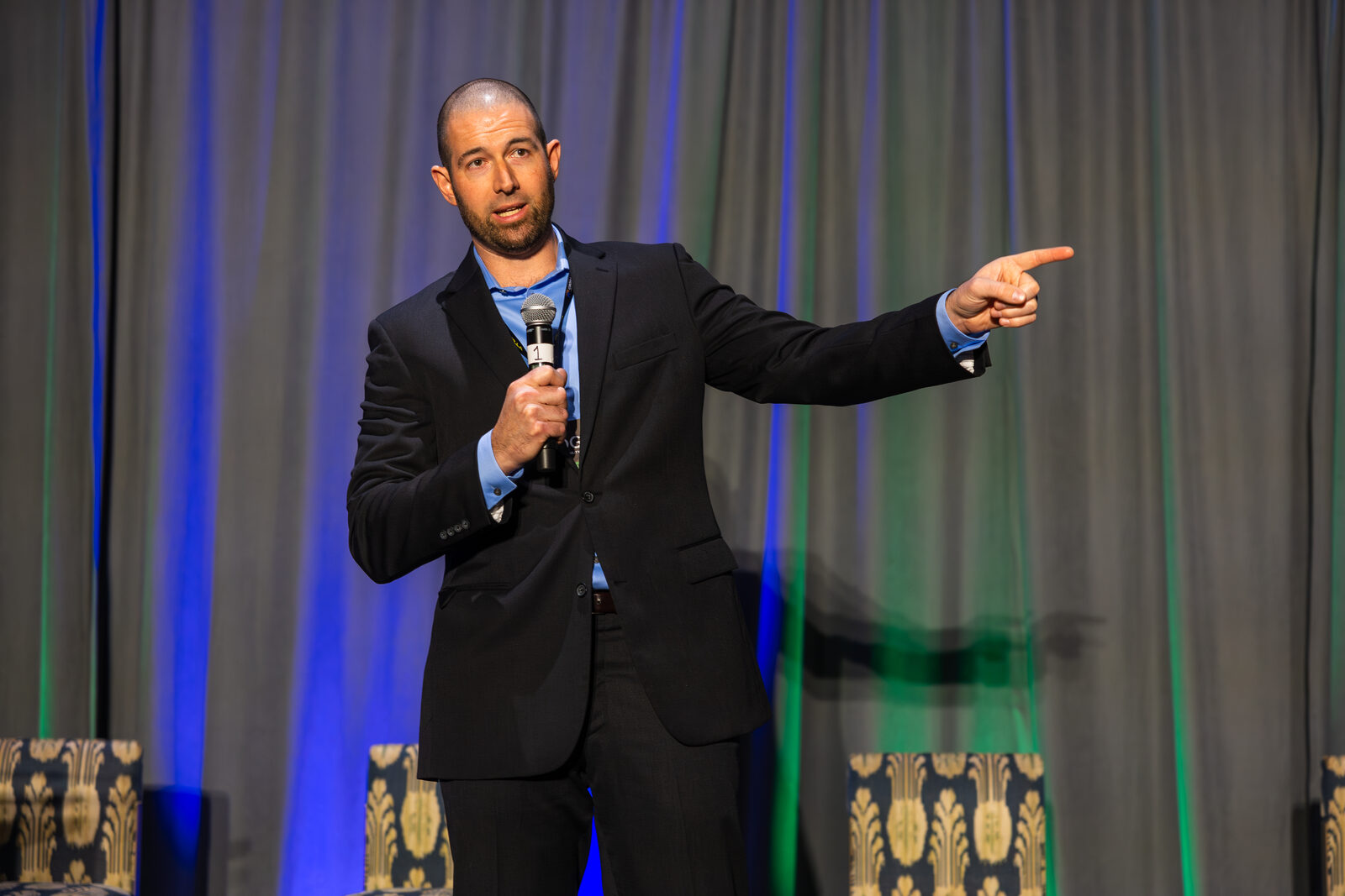 Dan Coomes-Barry on stage at AGOGE 2024, a microphone in one hand and the other pointing out into the audience.