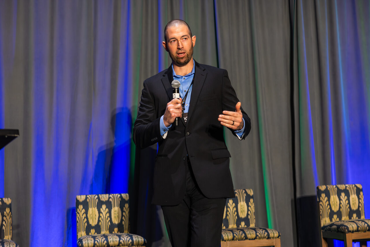Dan Coomes-Barry speaking into a handheld microphone on stage at AGOGE 2024.