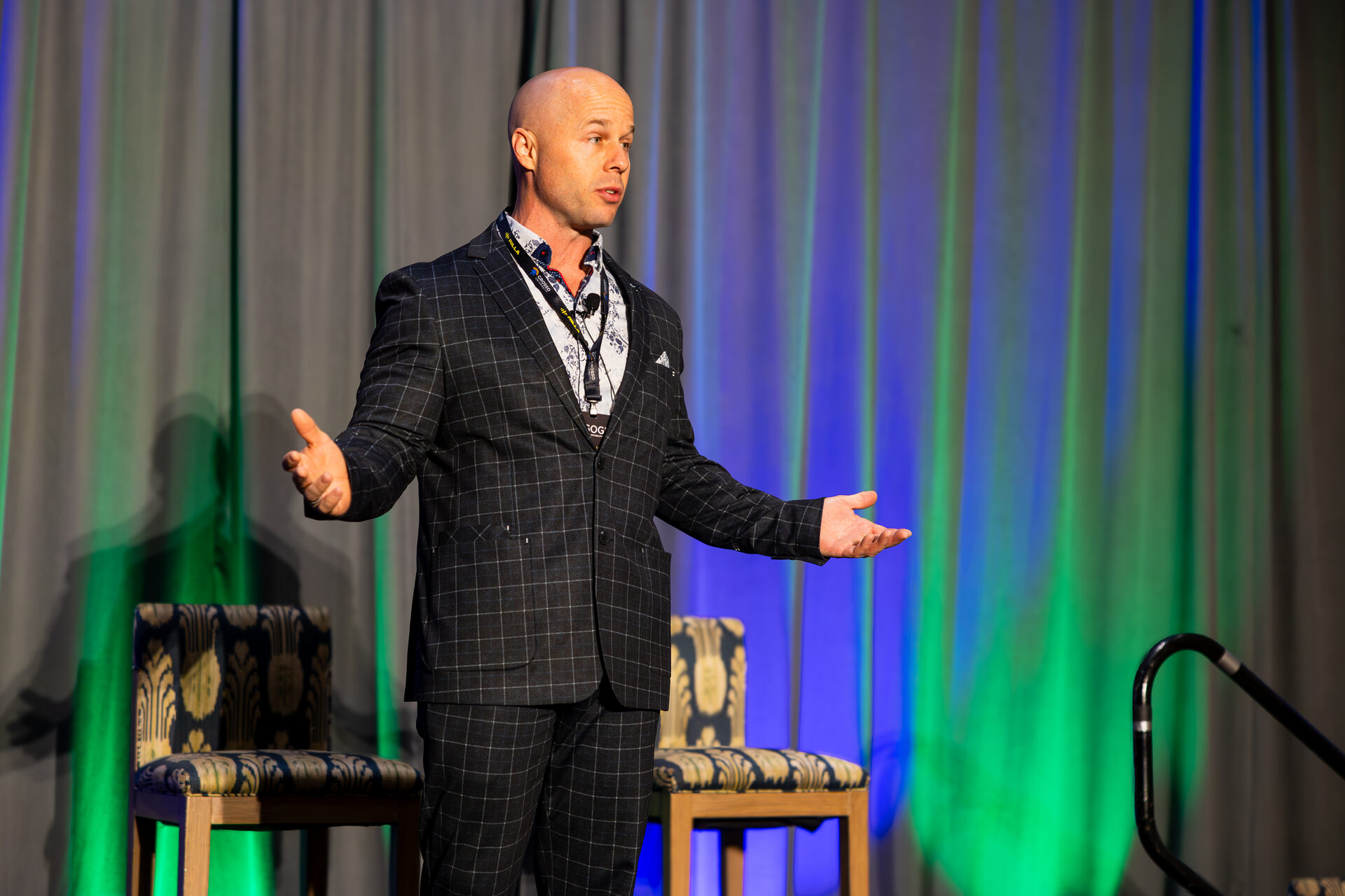 Dominic Caminata speaking on stage at AGOGE 2024 with both arms open, in front of blue and green uplit stage drapes.