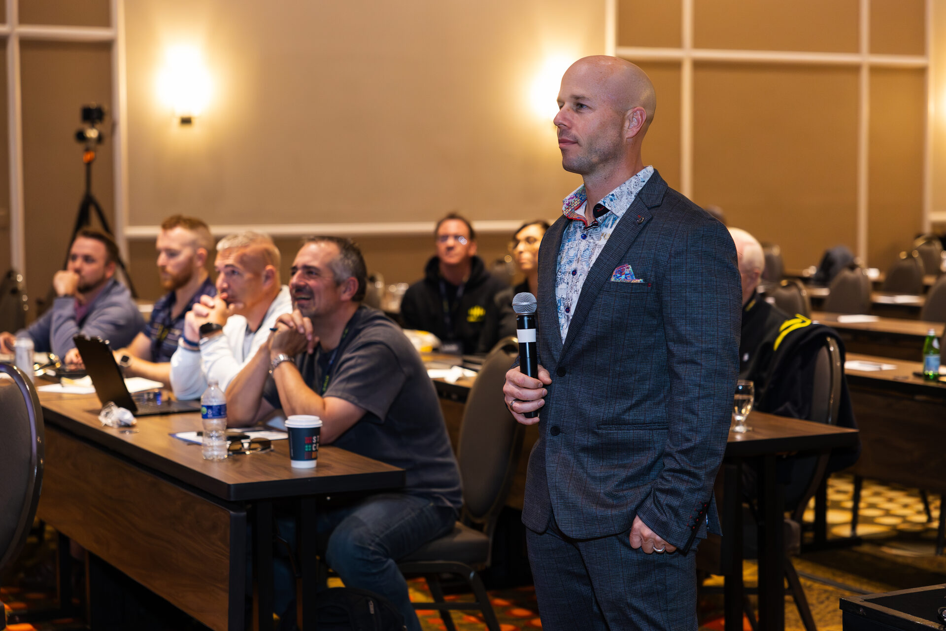 Dominic Caminata standing among the tables at an AGOGE 2024 breakout session, microphone in hand, as seated attendees turn to listen.