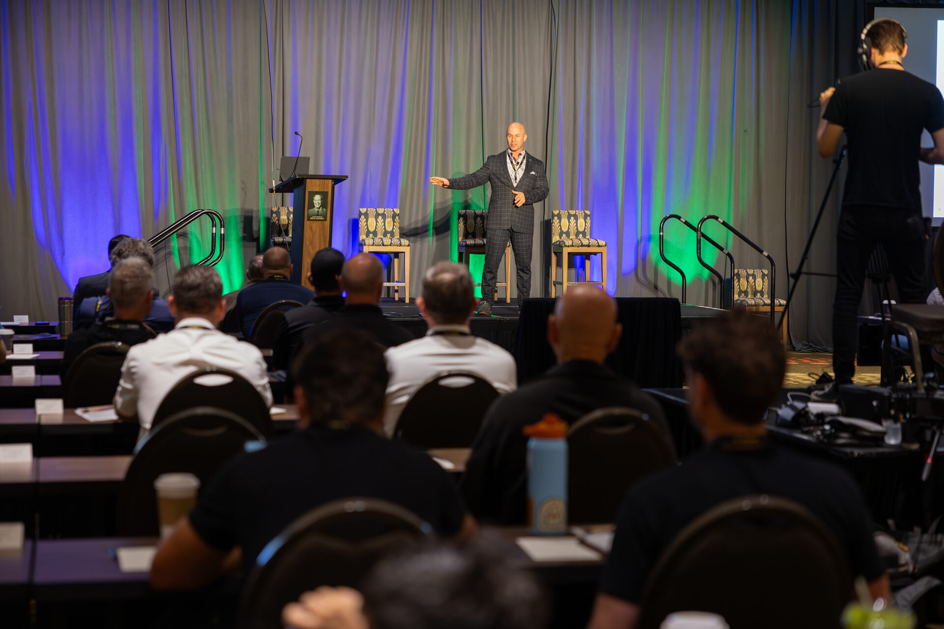 The AGOGE 2024 general session seen from the back of the ballroom: rows of seated attendees facing Dominic Caminata alone on a lit stage, with a videographer filming from the side.