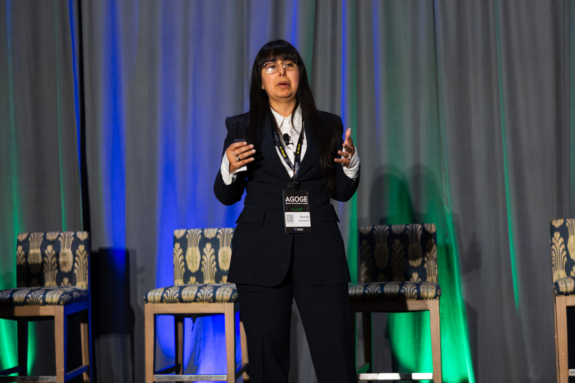 Wendy Harmeyer delivering her keynote on stage at AGOGE 2024, both hands raised as she speaks, wearing a keynote speaker badge.