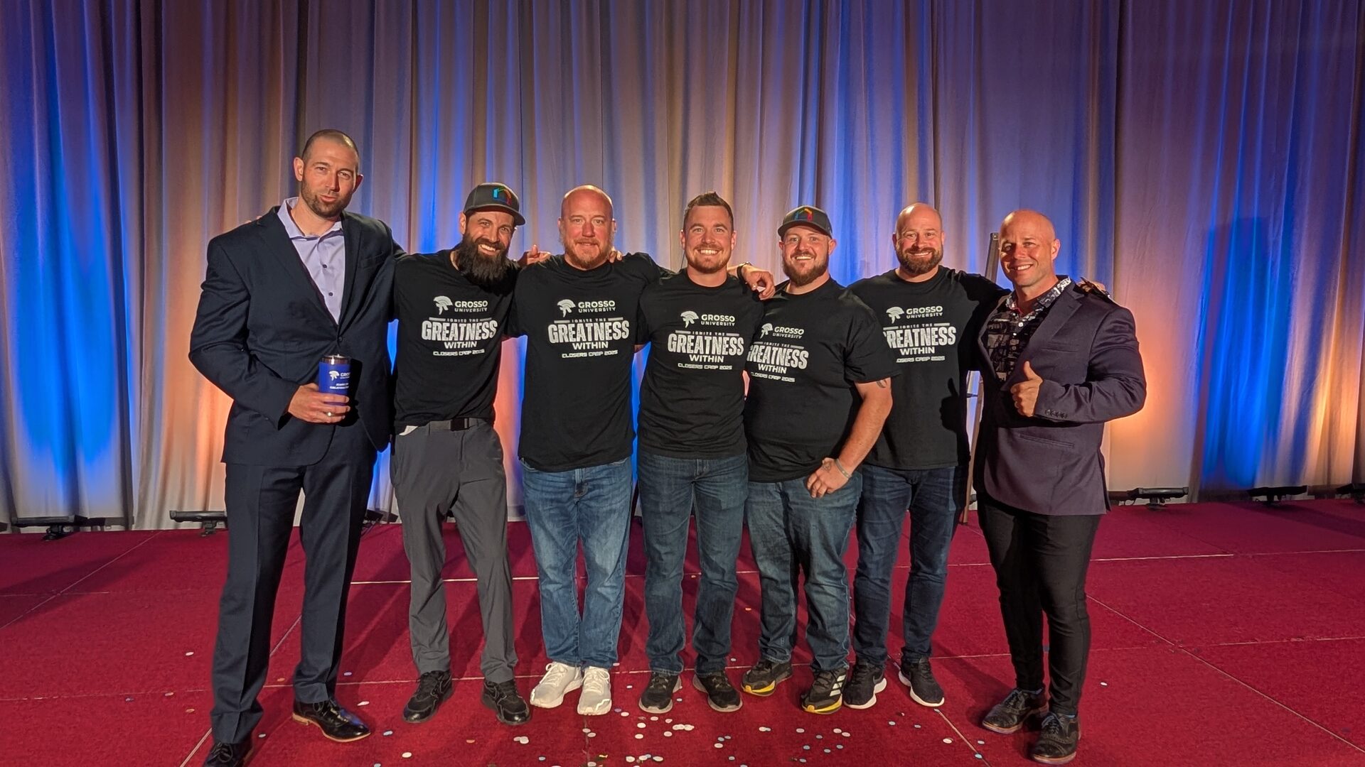 Seven Closers Camp 2025 attendees and staff standing shoulder to shoulder on the stage, most in Grosso University shirts.