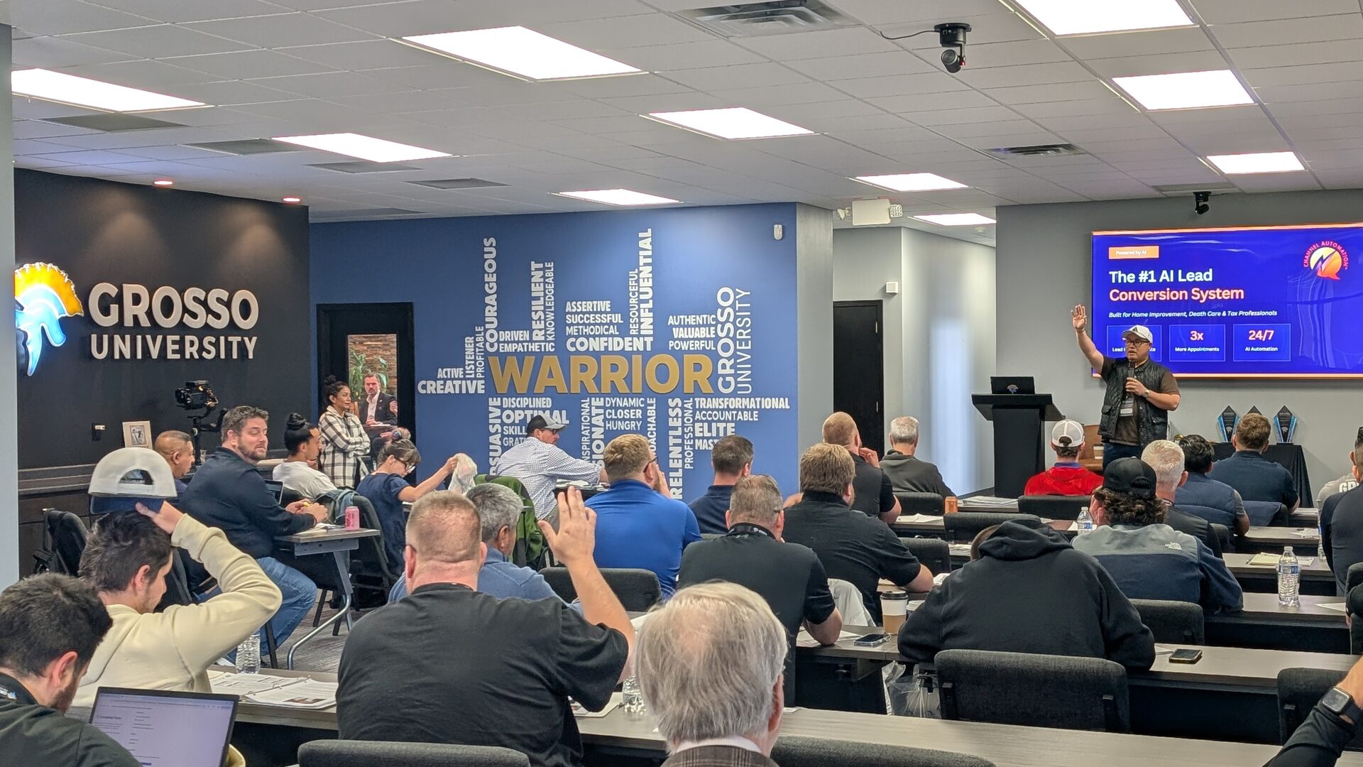 A wide view of the Grosso University training room during Closers Camp 2026, the illuminated lobby sign and Warrior word wall on the left and a presenter beside a slide titled "The #1 AI Lead Conversion System".