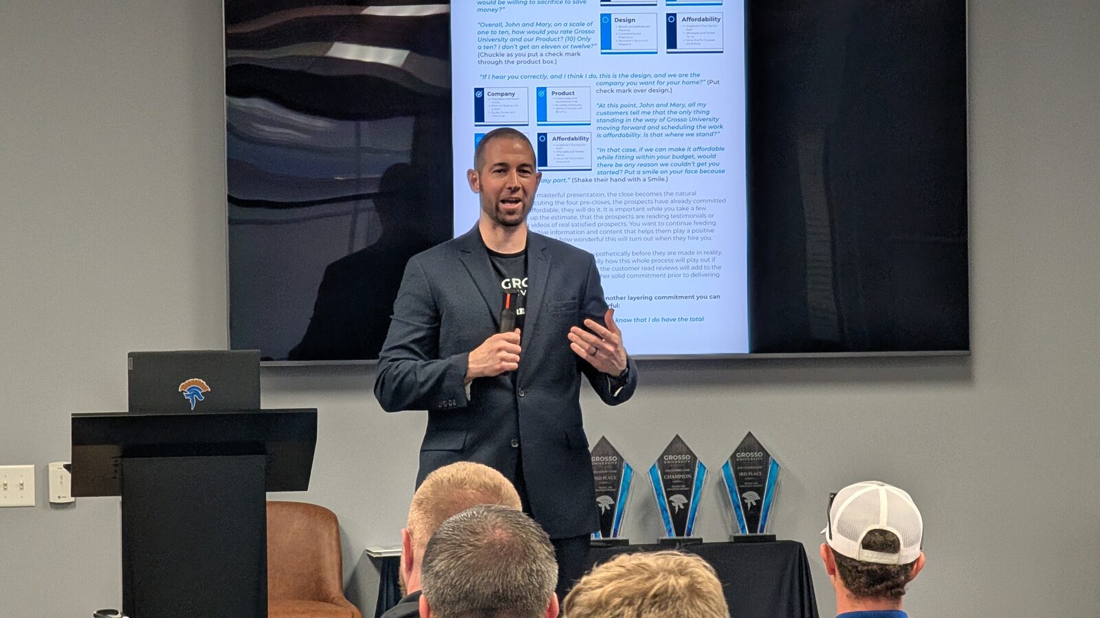 Dan Coomes-Barry presenting from a lectern at Closers Camp 2026, a slide behind him and three champion trophies on the table alongside.