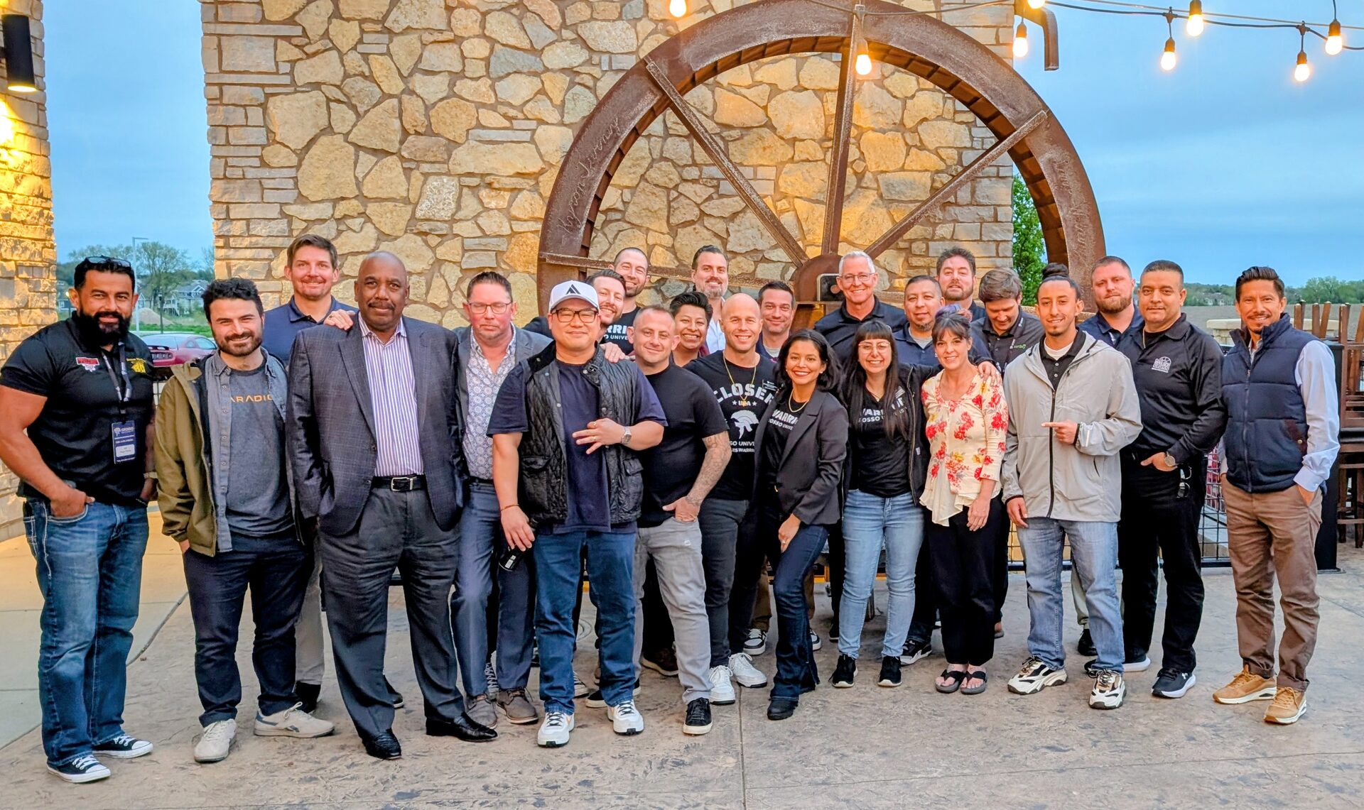 About twenty-five Closers Camp 2026 attendees and staff posed together outdoors at dusk in front of a stone wall and a large iron wagon wheel.