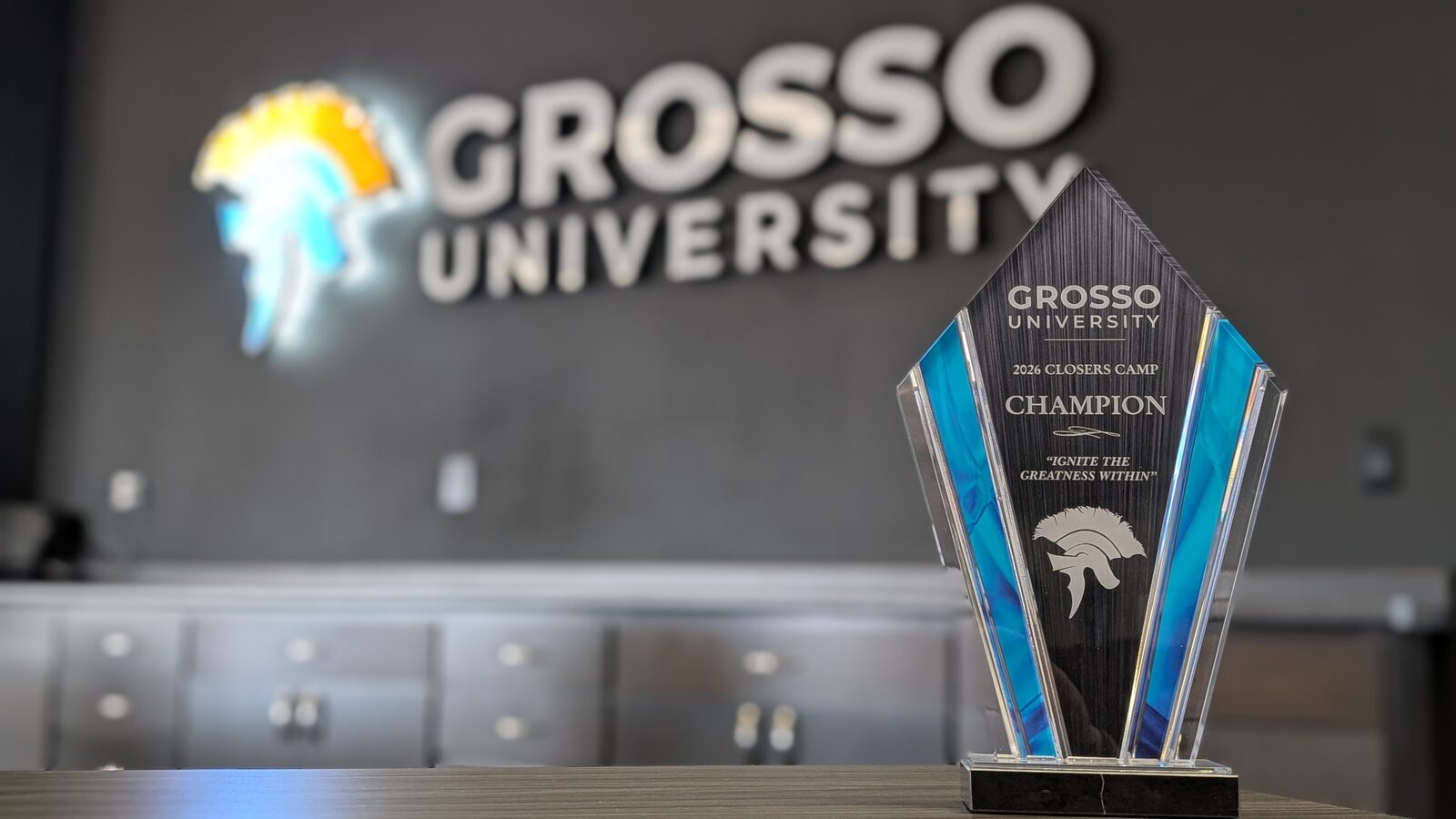 The 2026 Closers Camp Champion trophy, an engraved glass spartan helmet on a black base, on a table in front of the illuminated Grosso University lobby sign.