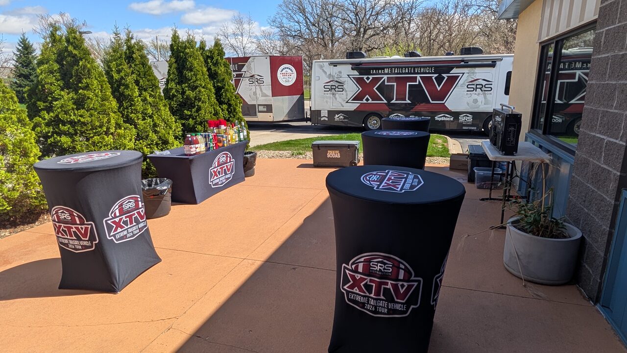 A wider view of the Extreme Tailgate Vehicle patio setup, the branded coach parked behind the cocktail tables.