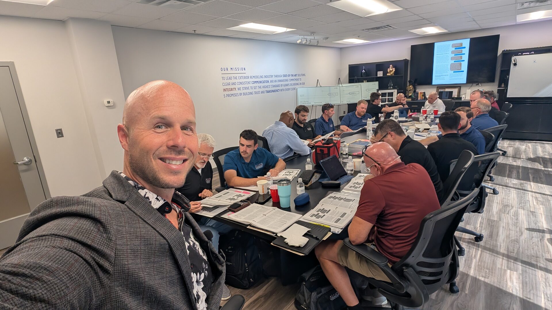Dominic Caminata in the foreground of a Grosso University conference room where roughly a dozen executives sit around a long table with printed workbooks, the company mission painted on the wall behind them.