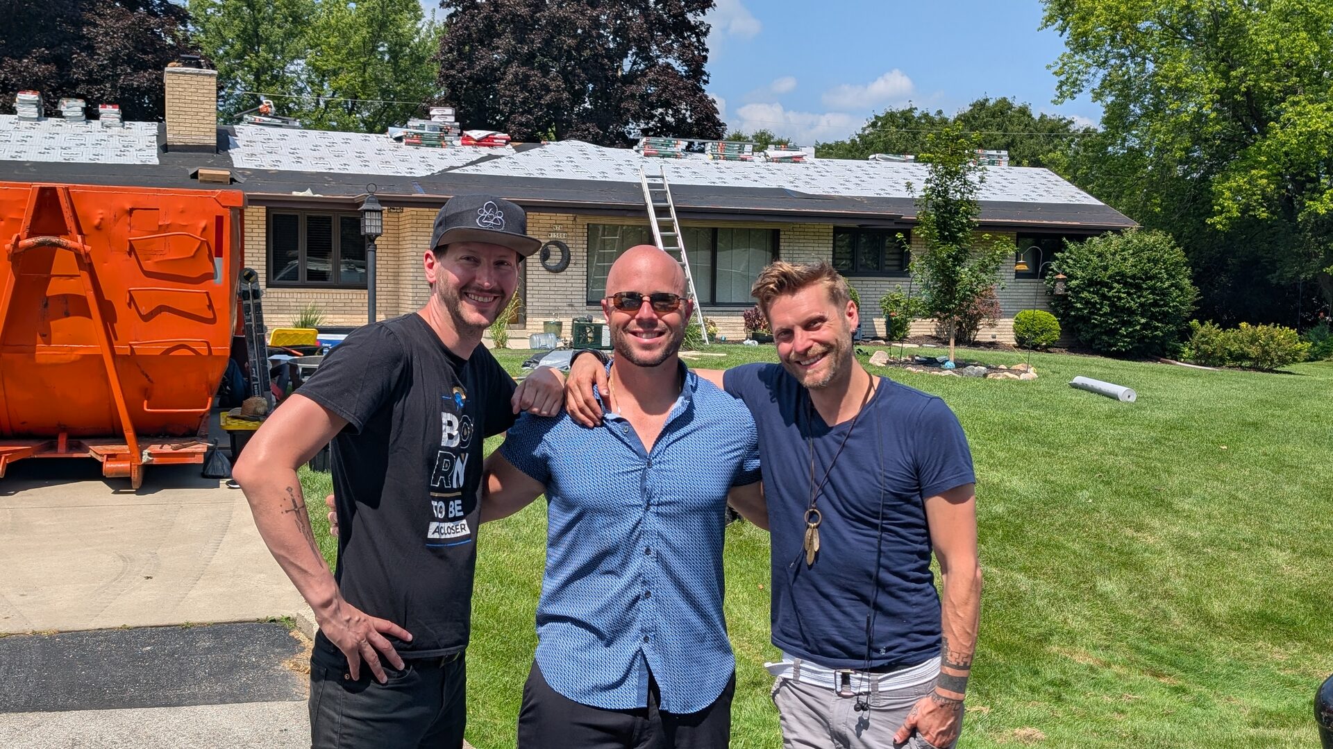 Brett Hill, Dominic Caminata and Brett Naples standing together on the lawn of a roofing job site, a dumpster and the stripped roof behind them.