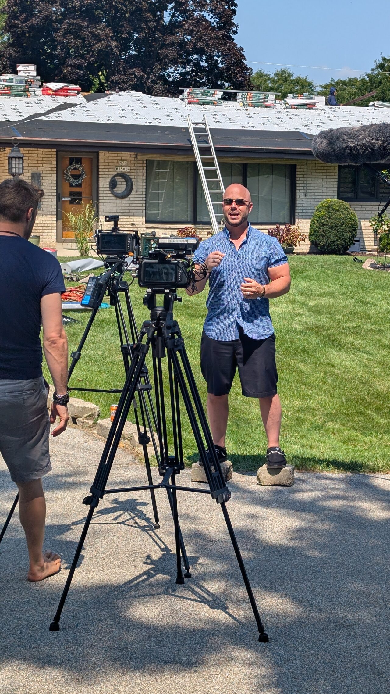 Dominic Caminata speaking to a camera on a tripod in the driveway of a house being re-roofed, a ladder against the eaves and bundles of shingles on the roof.