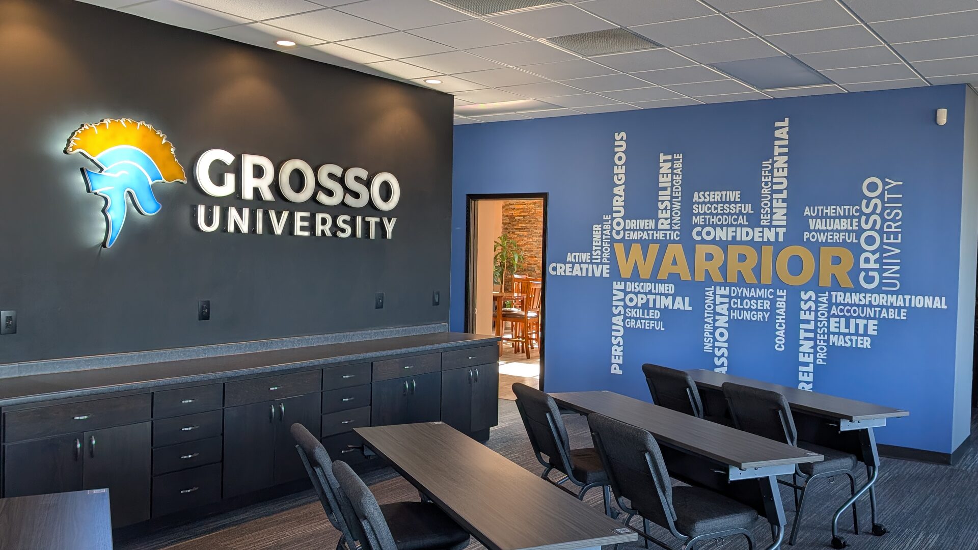 The Grosso University office interior: the illuminated spartan helmet and wordmark on a dark gray wall, the blue Warrior word wall beyond it, and training tables in the foreground.