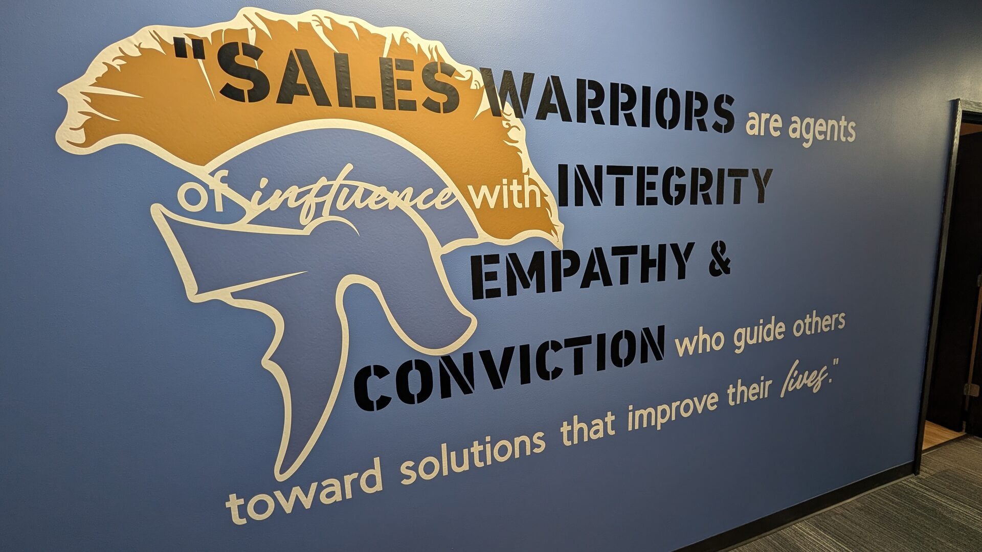 An office wall carrying the Grosso University spartan helmet and the line "Sales warriors are agents of influence with integrity, empathy and conviction who guide others toward solutions that improve their lives."