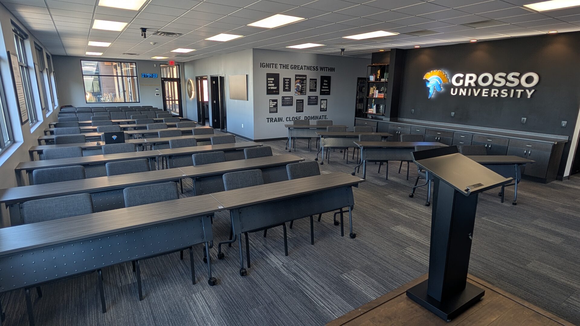 The empty Grosso University training room, rows of tables and chairs facing a lectern, the illuminated wordmark on the right and the "Ignite the greatness within" poster wall at the back.