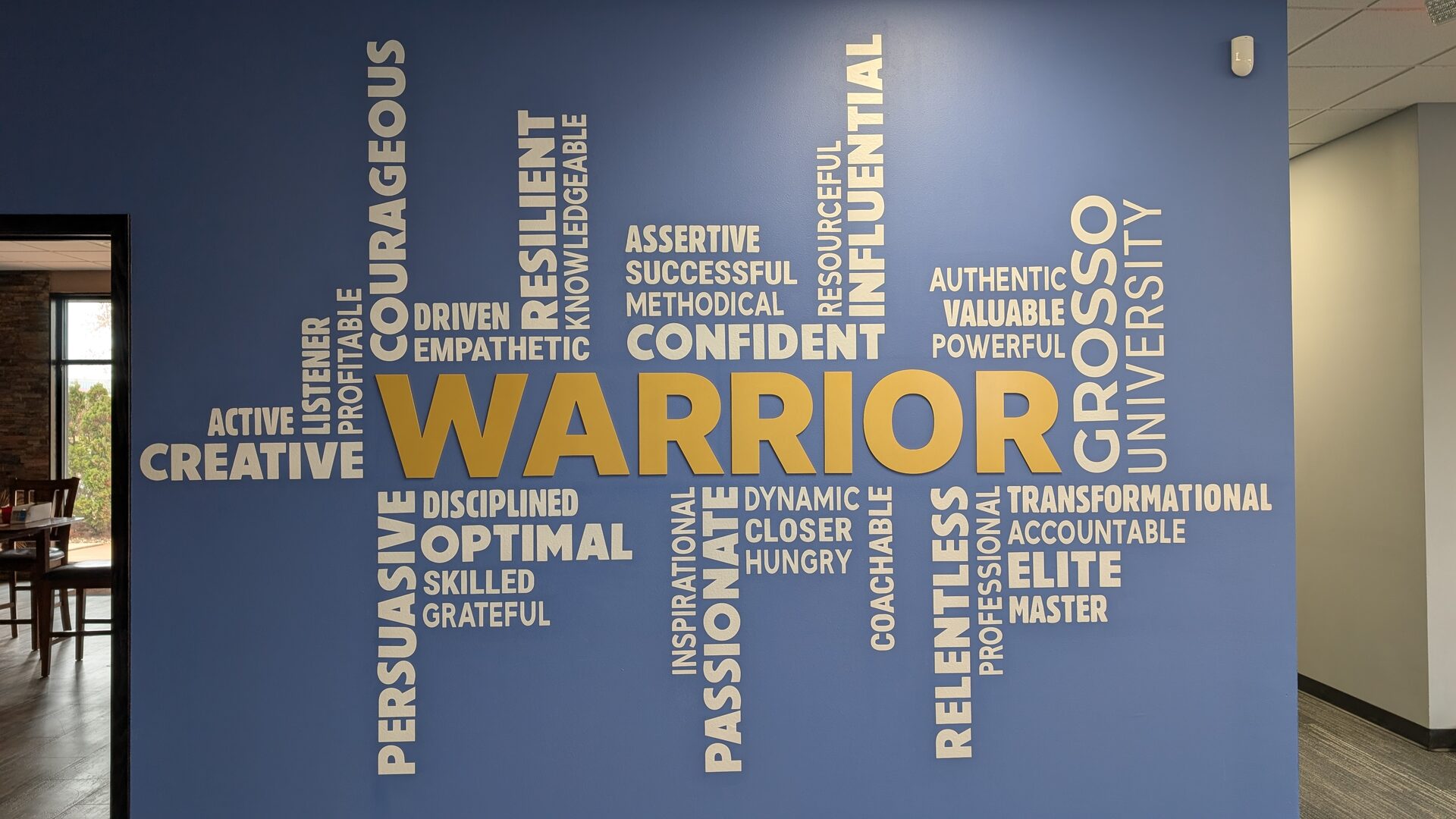 A blue office wall covered in a word cloud built around the word "Warrior" in gold, surrounded by traits including courageous, disciplined, relentless and coachable.