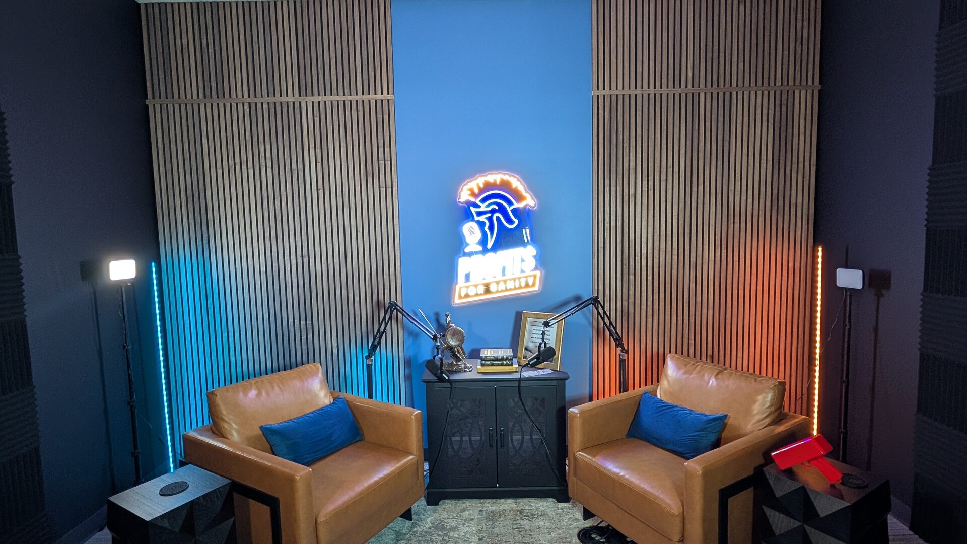 A wide view of the Profits for Sanity podcast set showing the two armchairs, the neon sign, slatted wood walls and studio lighting on either side.