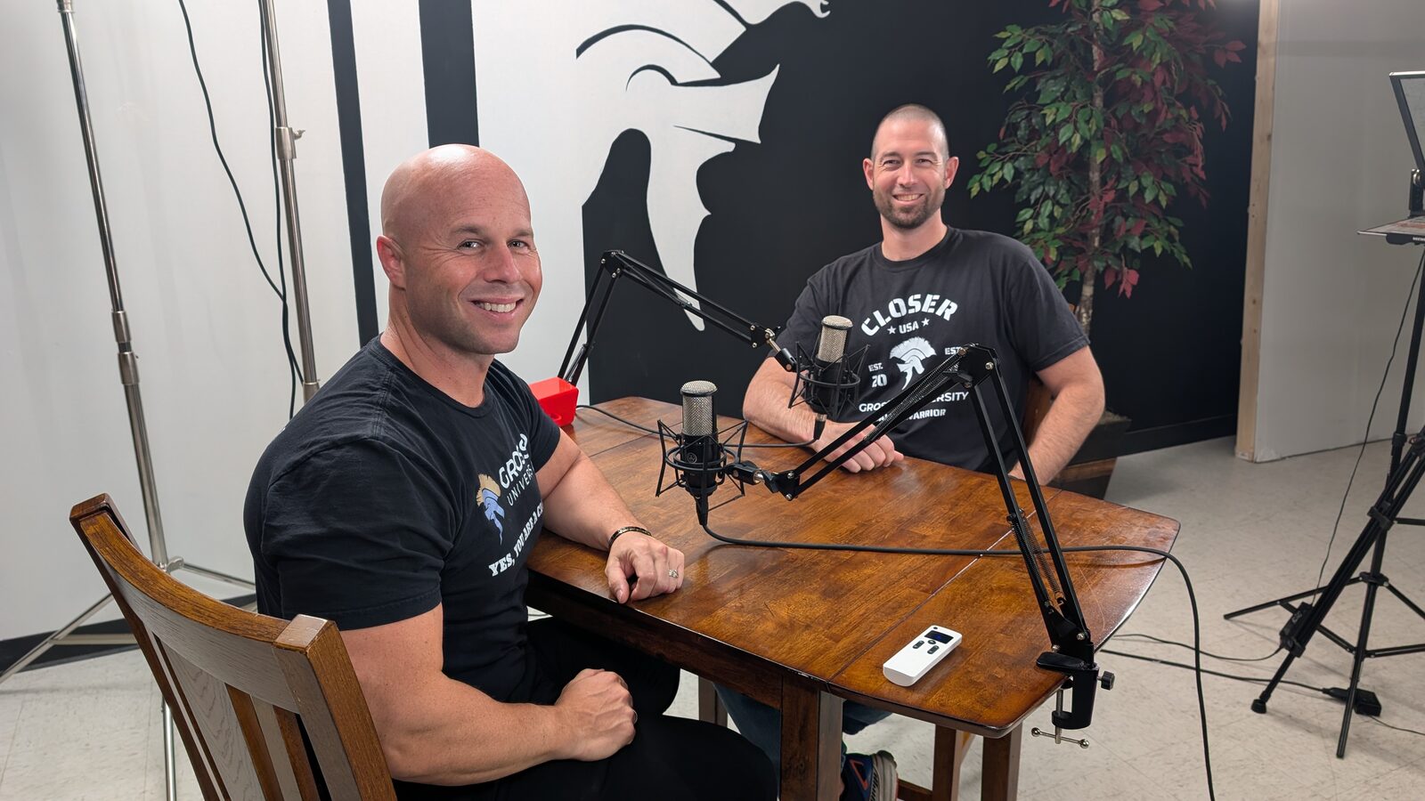 Two Grosso University hosts seated at a wooden table with studio microphones, in front of a wall painted with the Grosso University spartan helmet.